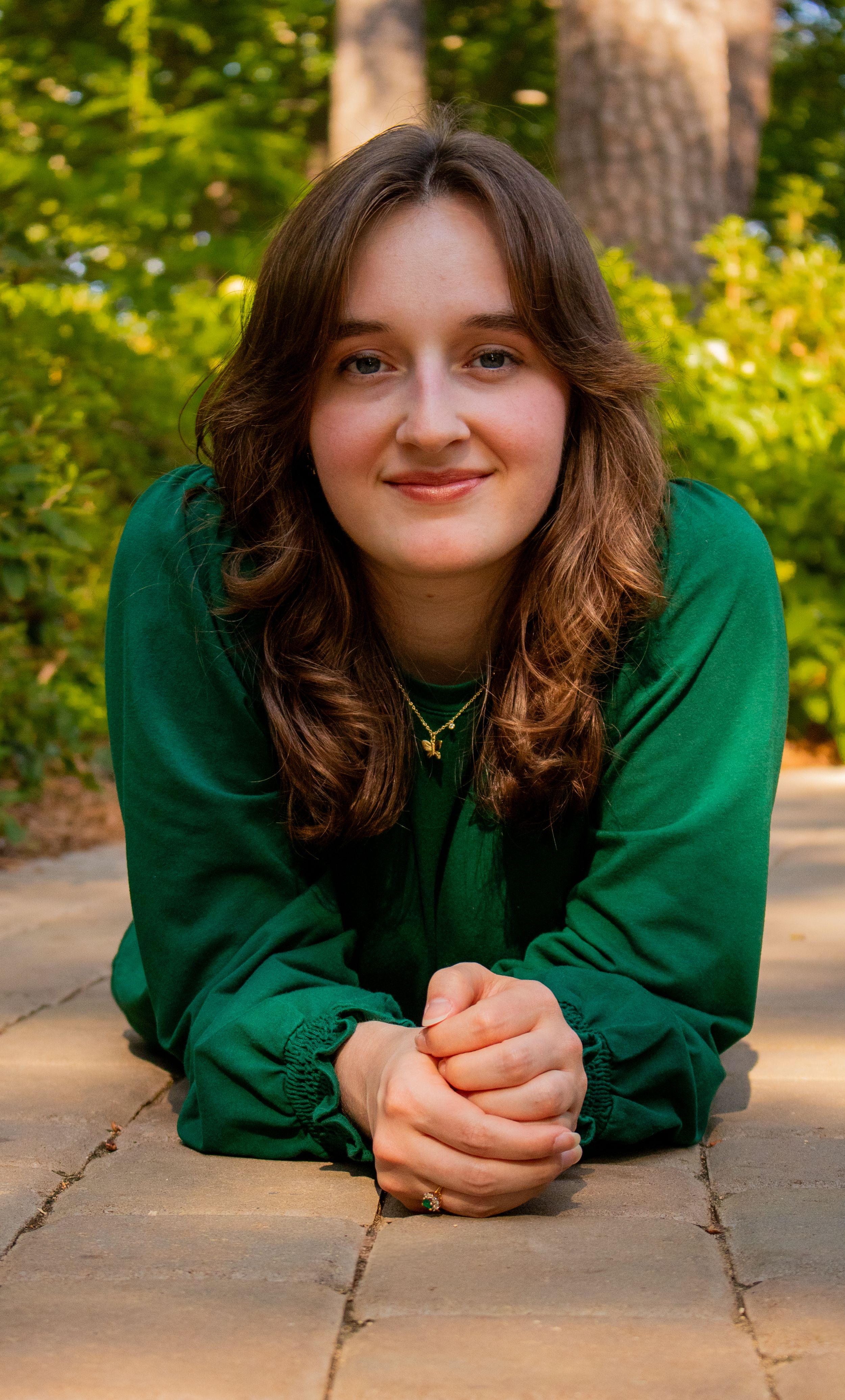 Young woman in green hoodie lying on brick pavement, smiling outdoors with lush green trees in background.