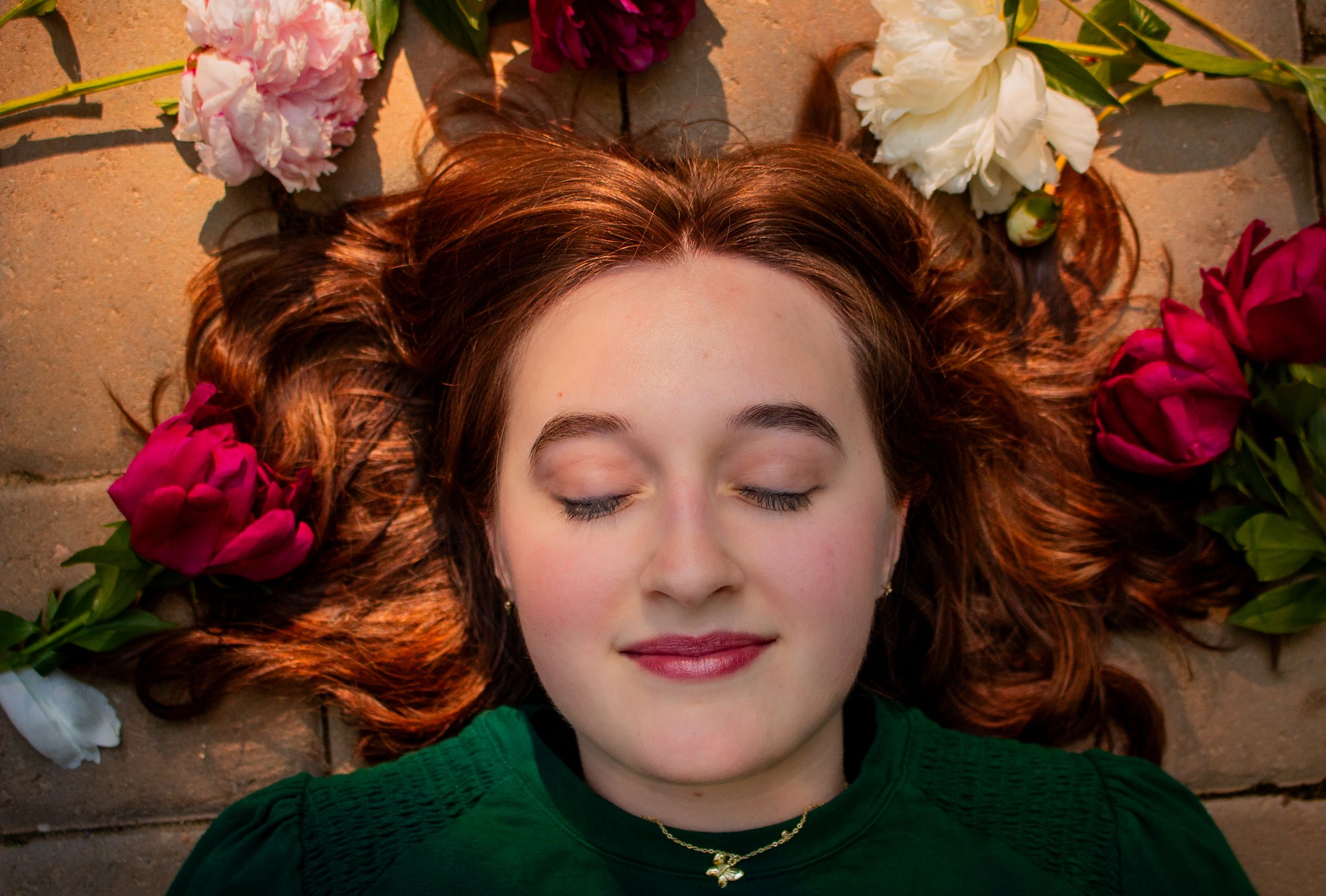 Young woman with red hair lying among colorful peonies, eyes closed, wearing a green top and gold necklace.
