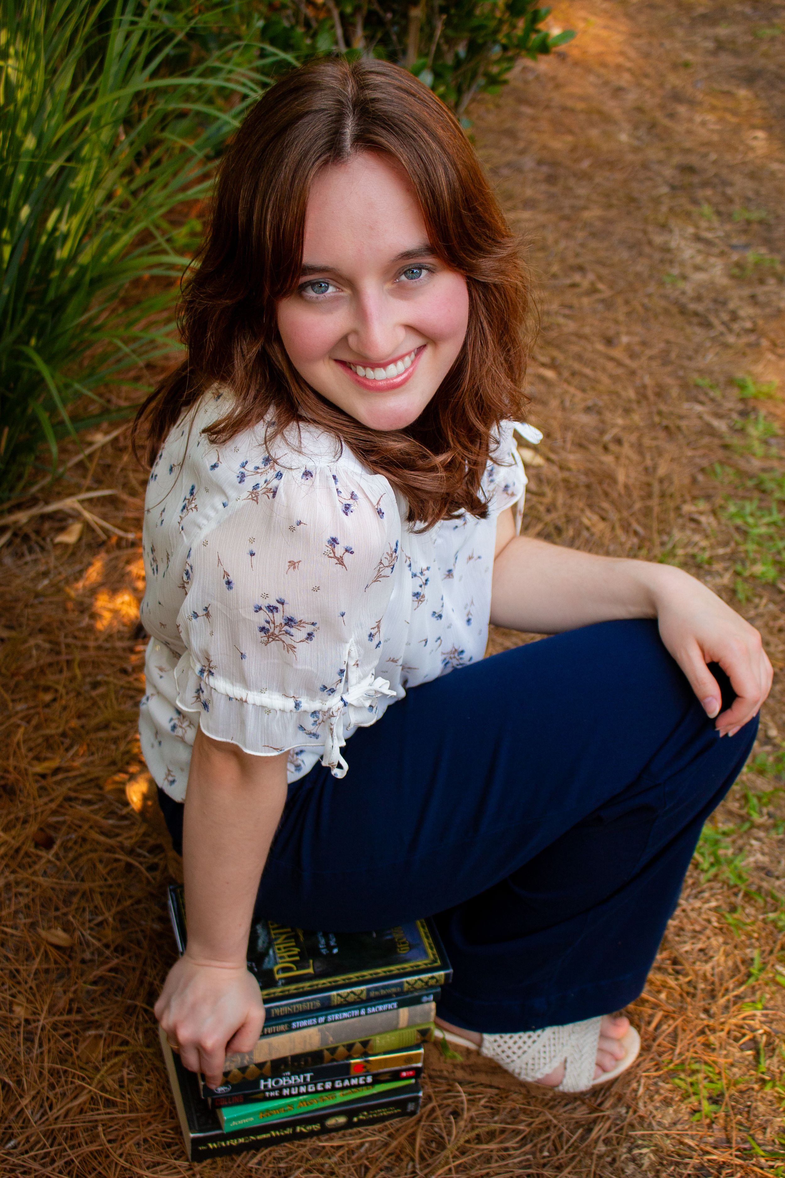 Young woman with auburn hair smiling outdoors, sitting on books in navy pants and floral blouse.