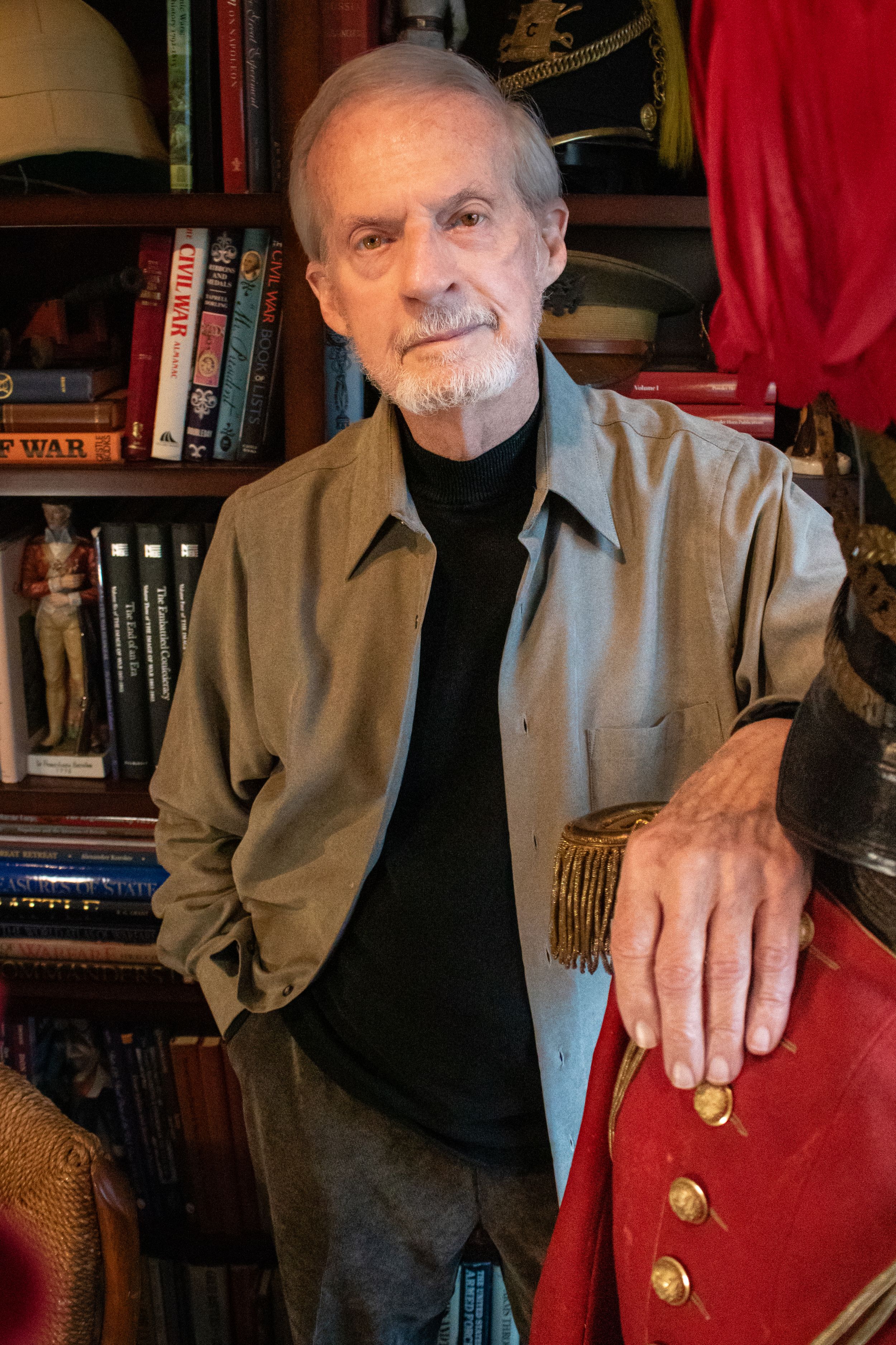 Elderly man with white beard poses in home library with military artifacts, books, and collectibles on shelves.