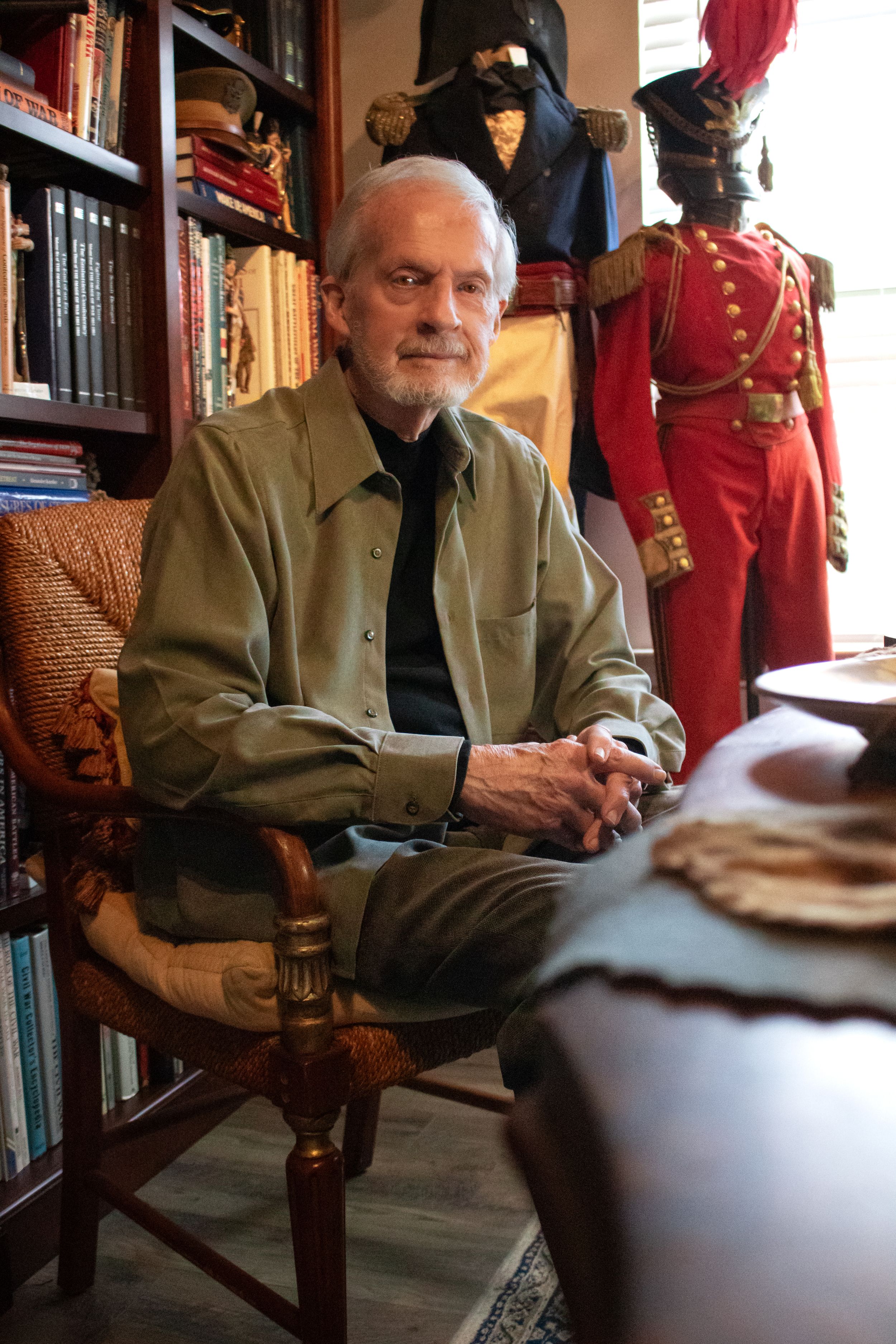Elderly man with white beard sits smiling in chair beside bookshelf and vintage military uniforms on display.