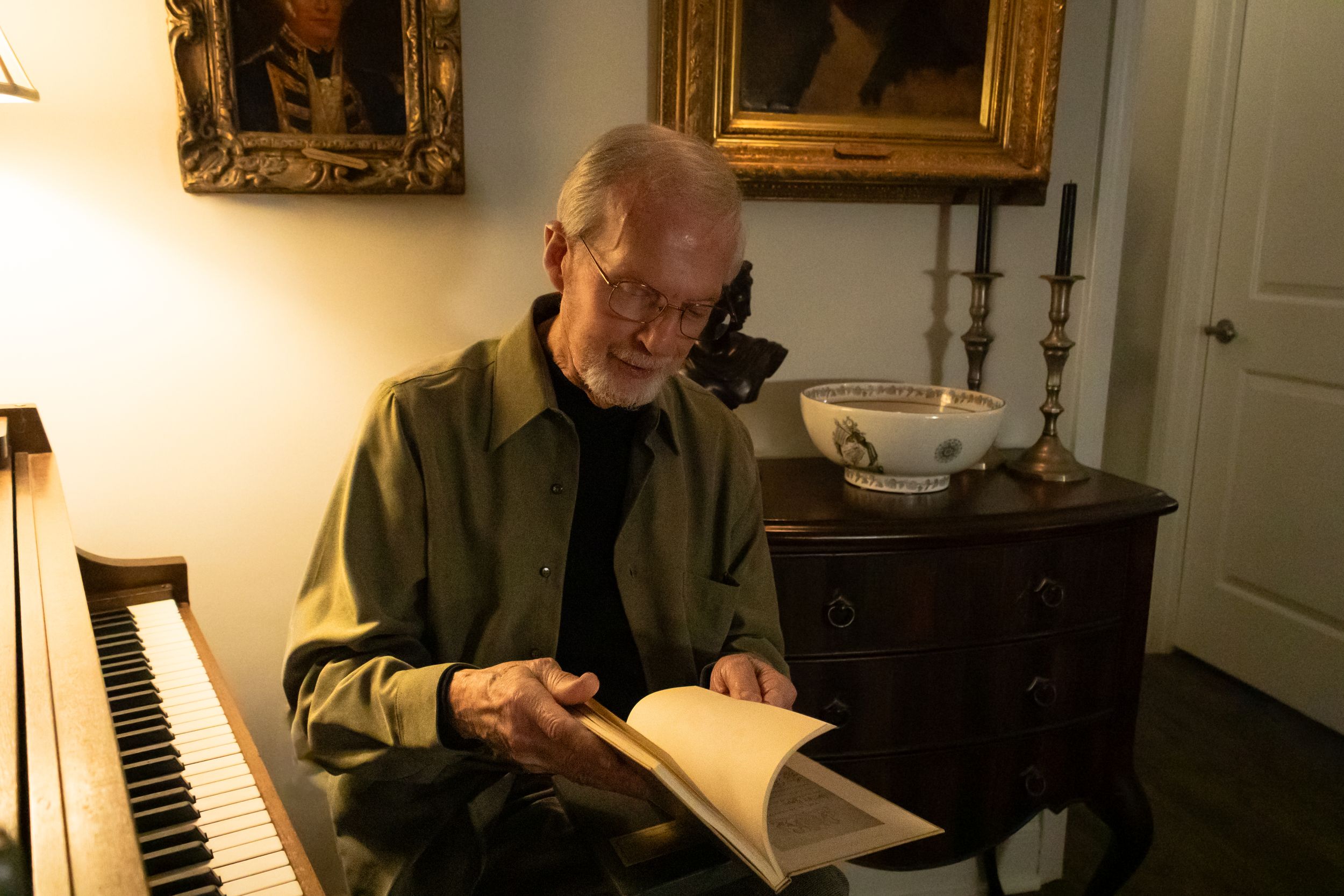 Elderly man reading a book near a piano in a warmly lit room with artwork and antique furniture.