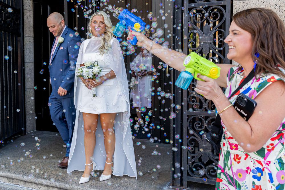 Bride and groom exit building as guest shoots bubbles with toy gun at outdoor wedding celebration.