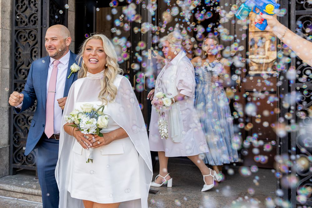 Bride in white mini dress holding bouquet exits church amid bubbles with groom in pink tie and guests behind.