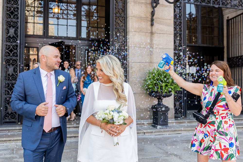 Bride and groom exit church as guest throws confetti during sunny outdoor wedding celebration.