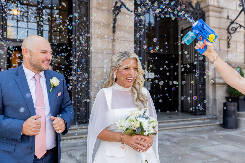 Happy newlyweds celebrate outside church with bubbles, bride holding bouquet, groom in blue suit with pink tie.