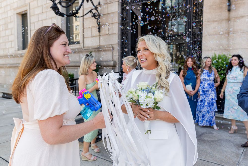 Bride in white cape gown holds bouquet while guests celebrate with confetti outside elegant venue.