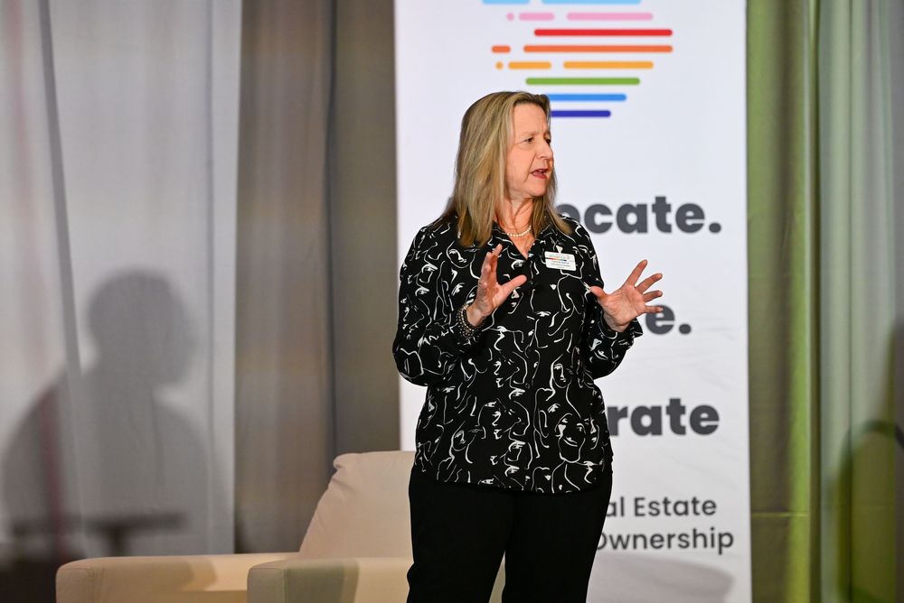 Woman in black patterned blouse speaks at real estate ownership advocacy event near colorful branded banner.