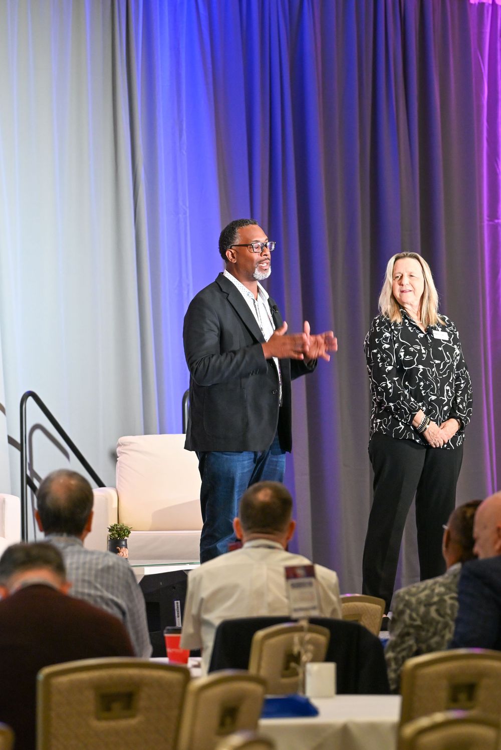 Two speakers present on stage at a professional conference, engaging an audience seated at round tables.