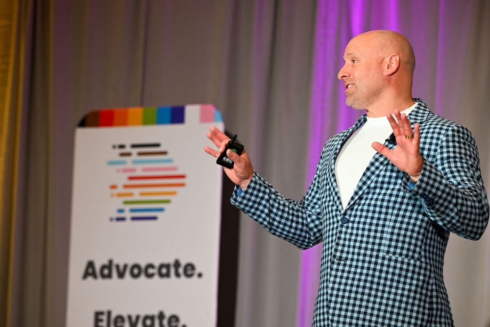 Bald man in checkered blazer speaking at podium with Advocate Elevate sign at corporate event.
