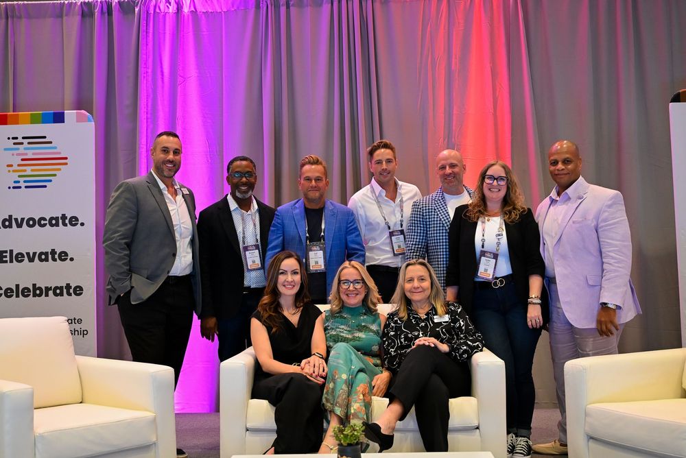 Group of ten diverse professionals posing together at a corporate event with colorful backdrop and white chairs.