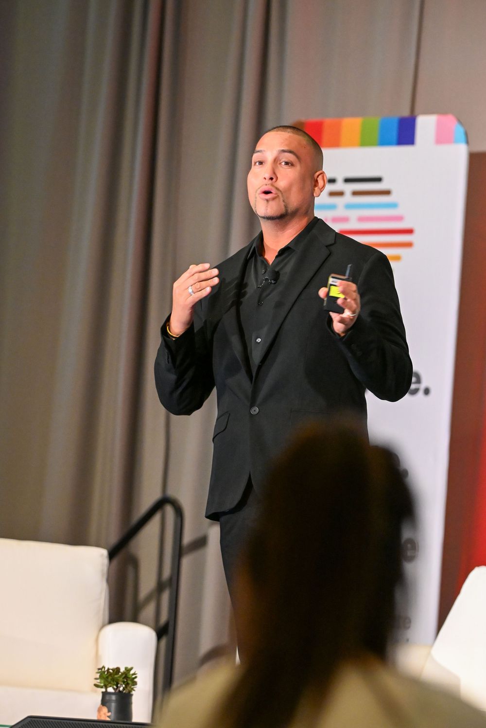 A dynamic speaker in a black suit presents on stage at a colorful corporate event.