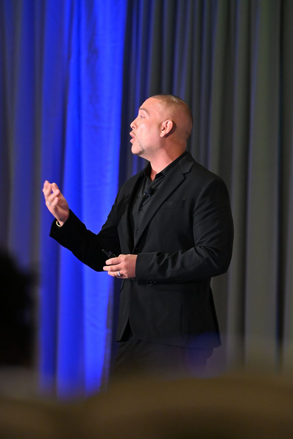 Bald man in black suit gesturing while speaking on stage with blue curtain backdrop.
