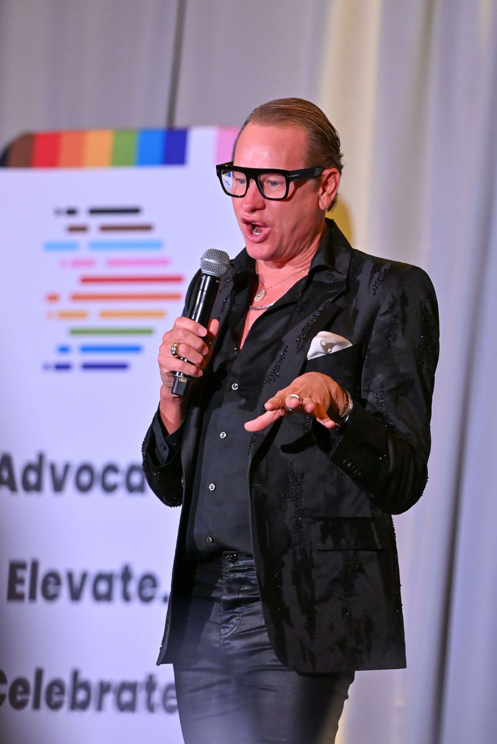Man in black blazer and glasses speaks at microphone at an Advocate, Elevate, Celebrate event.