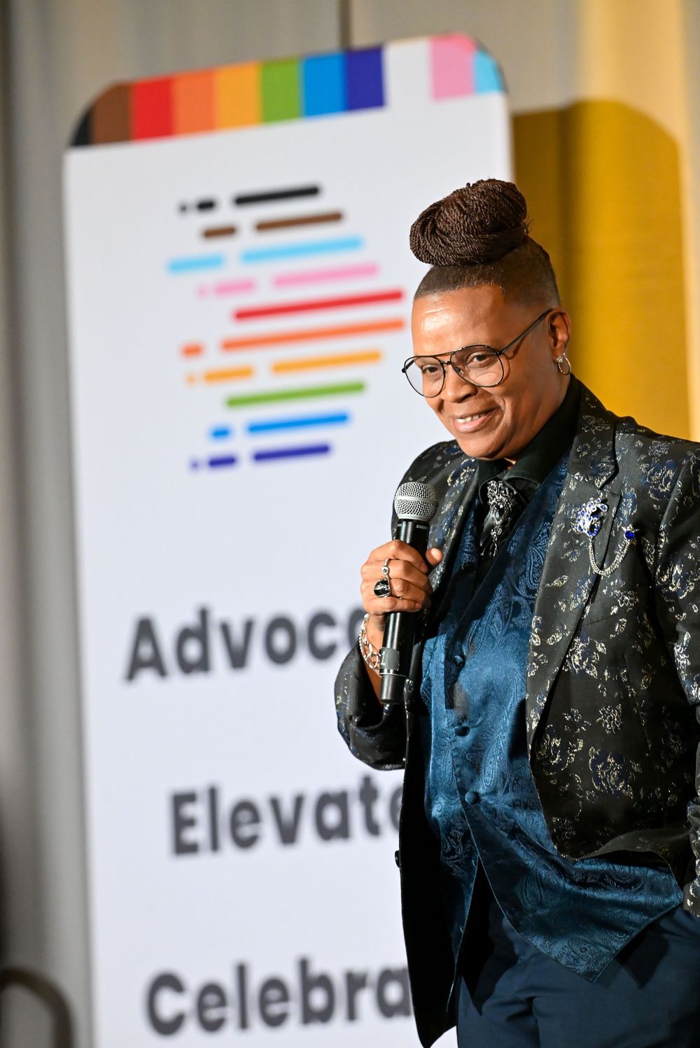 Person speaking at LGBTQ+ advocacy event, holding microphone, wearing teal blazer, standing near colorful pride banner.