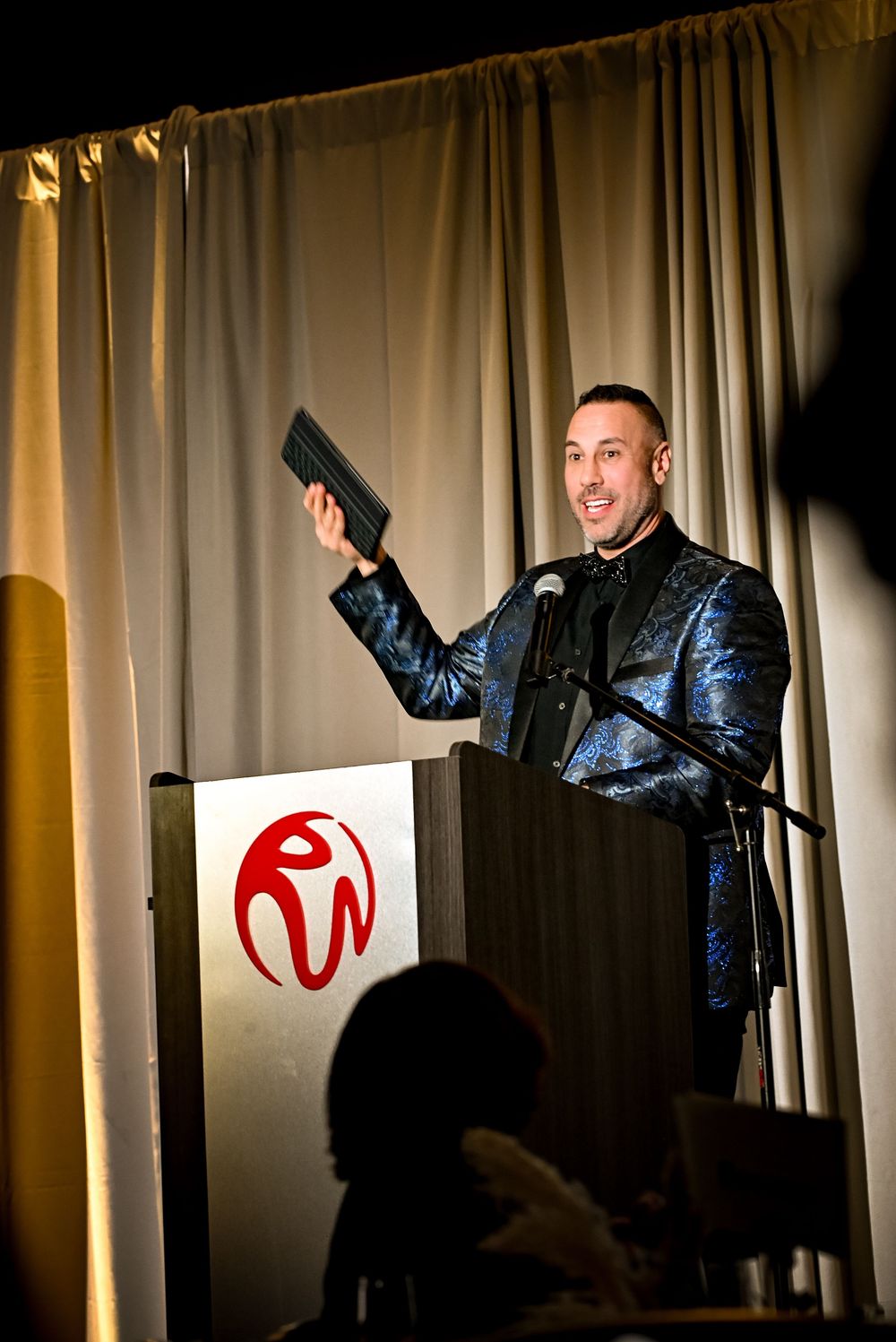 Man in blue sequin blazer speaks at podium with red logo, holding item aloft at formal event.