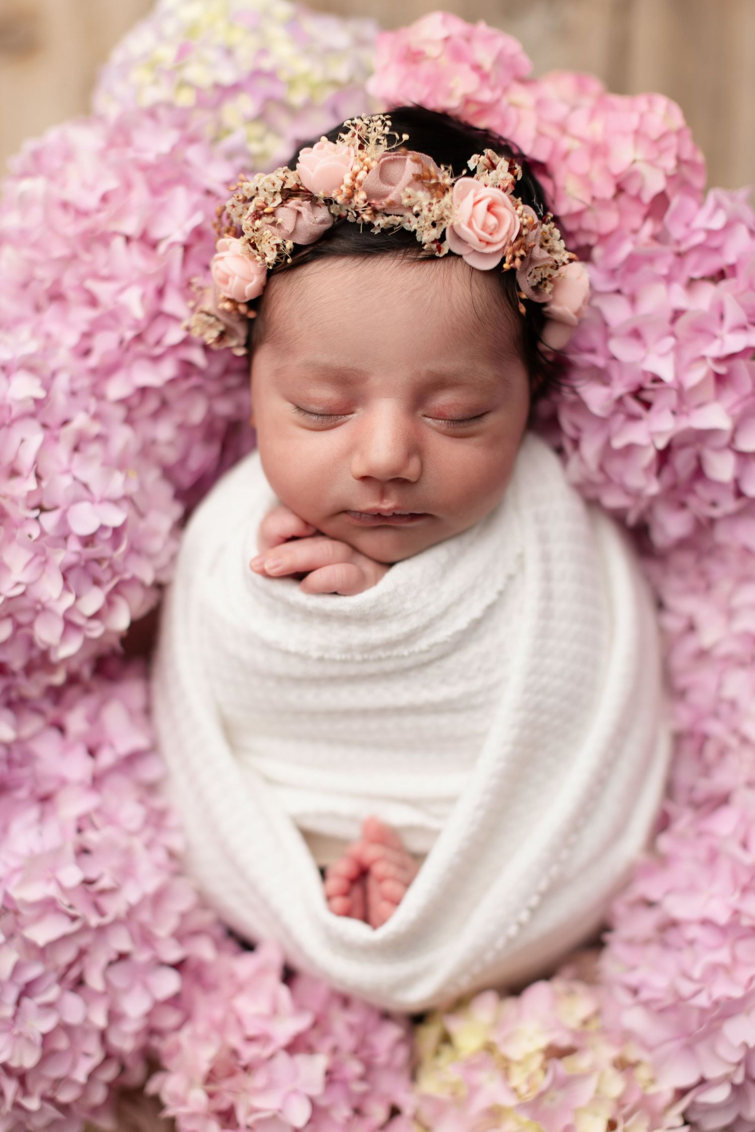 Sleeping newborn baby swaddled in white wrap, wearing a pink floral crown, surrounded by pink hydrangea flowers.