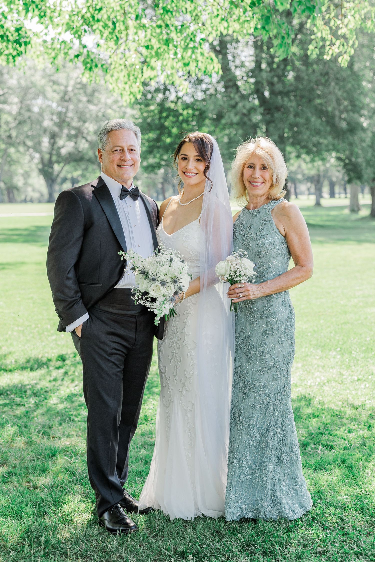 Bride in white gown with parents in formal attire posing outdoors in a lush green park on wedding day.