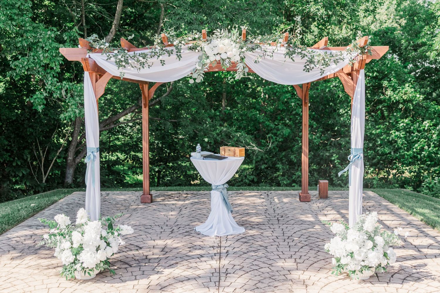 Elegant outdoor wedding ceremony arch with white draping, floral accents, and white flower arrangements on a brick patio.