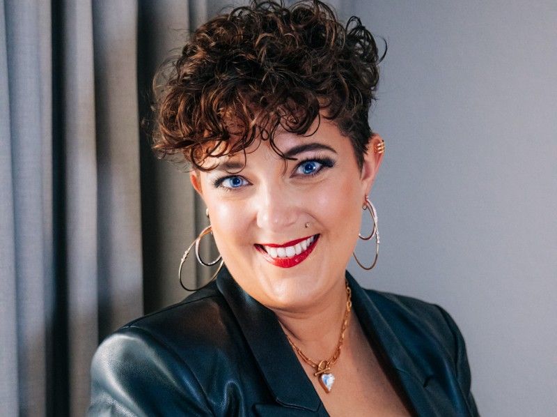 Woman with curly hair in black leather blazer and red top smiles confidently, wearing gold hoop earrings and necklace.