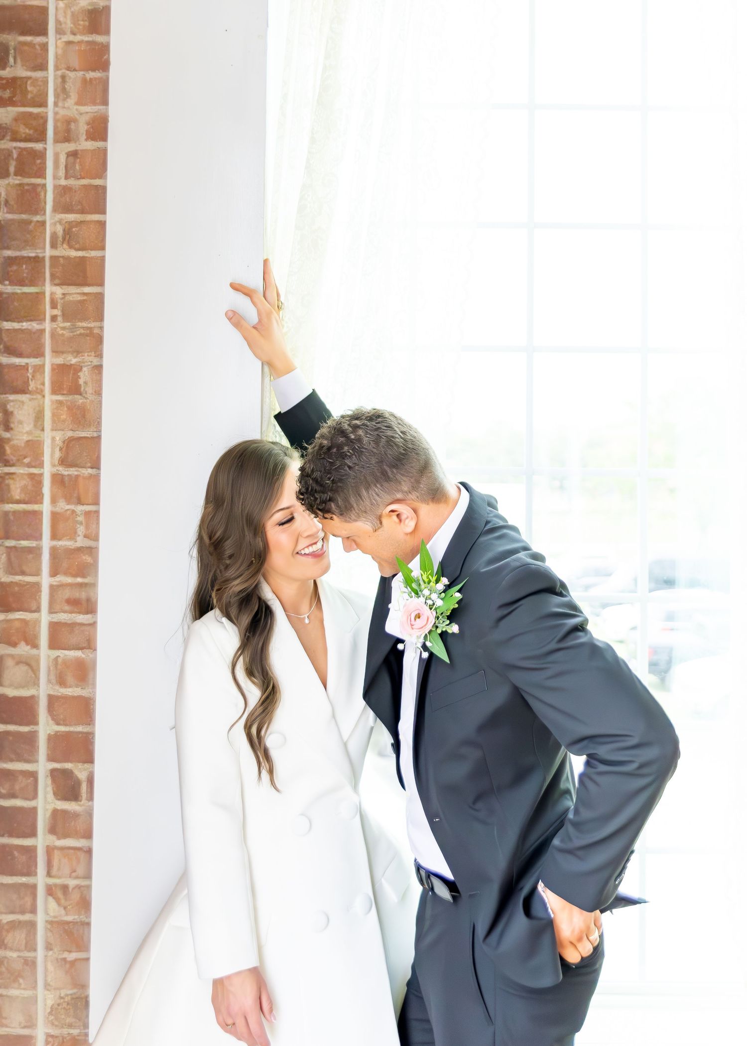 Bride in white blazer dress and groom in gray suit sharing a romantic moment by a brick wall and window.
