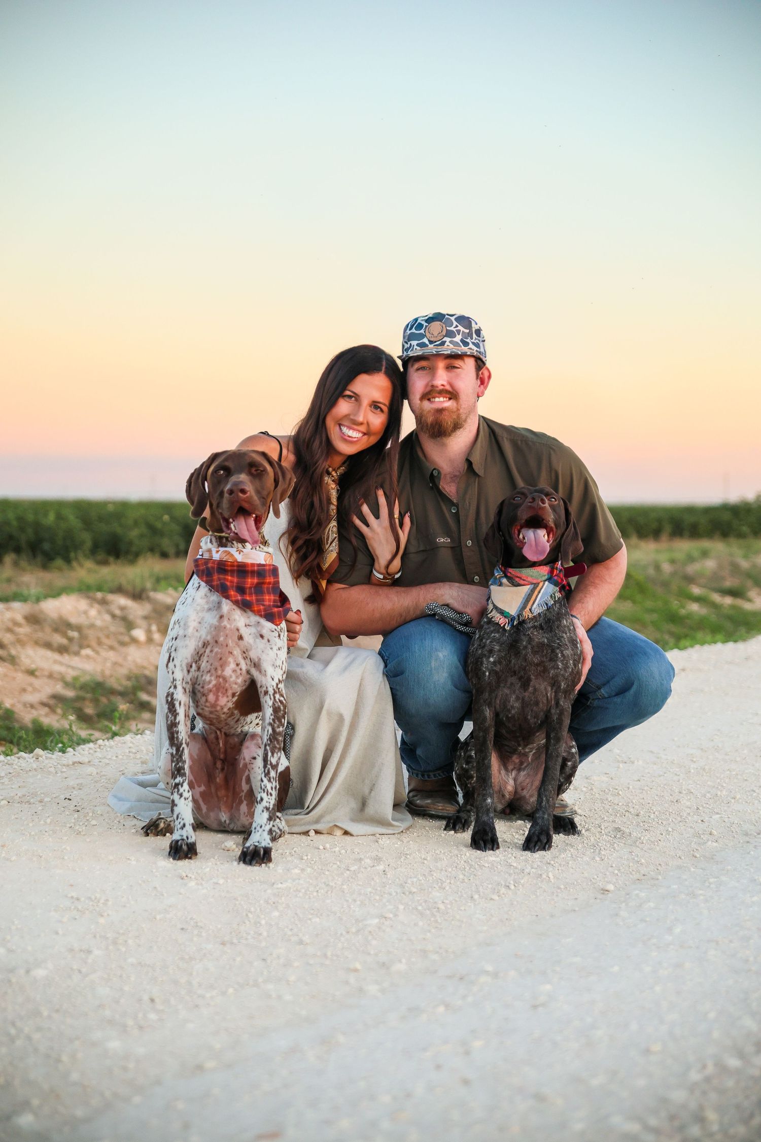 Happy couple crouching with two German Shorthaired Pointer dogs on a gravel path at sunset.