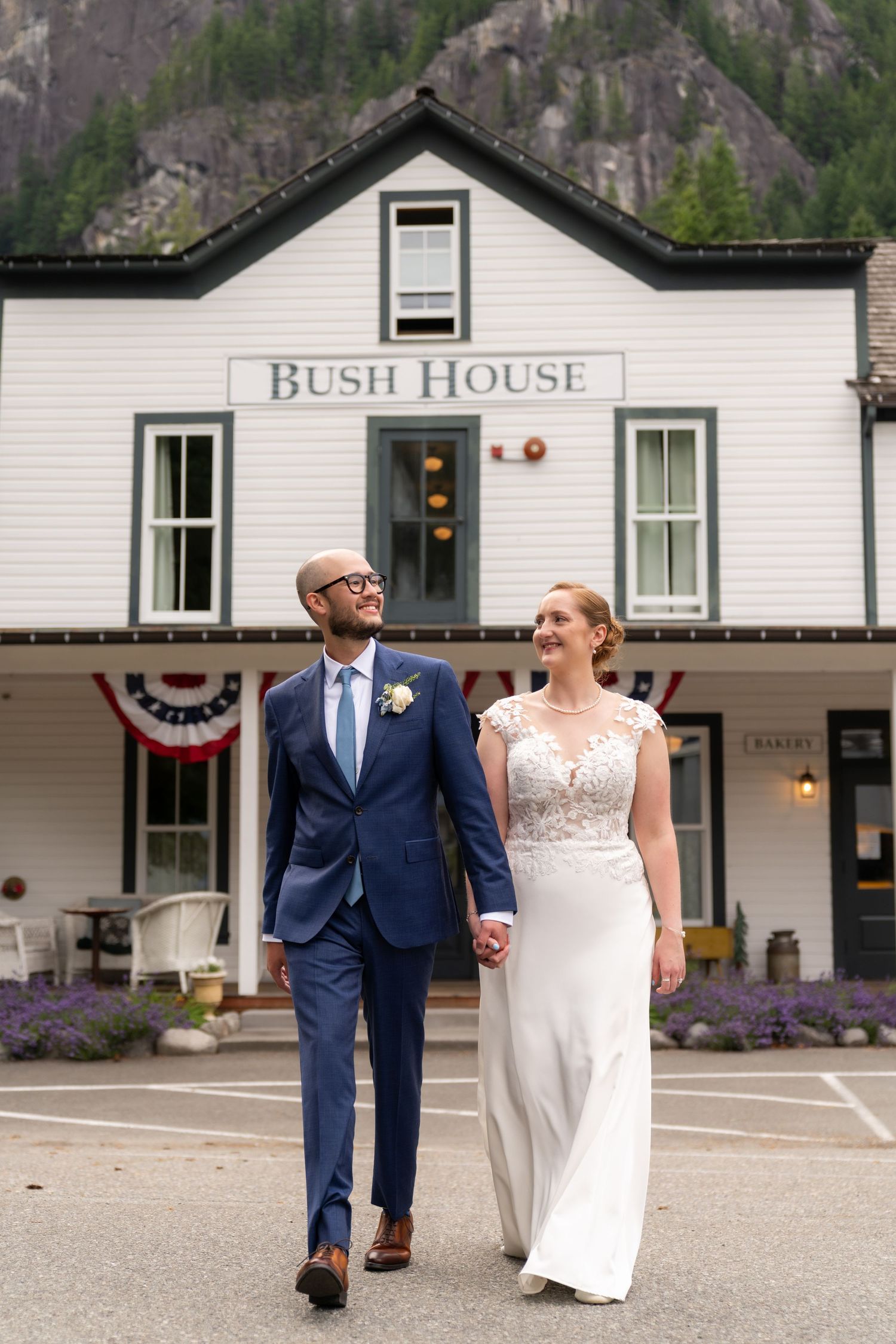 A bride and groom hold hands walking in front of the historic Bush House building with mountain backdrop.