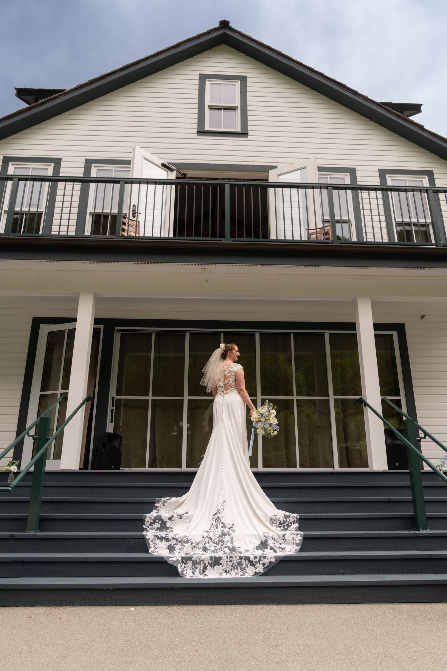 Bride in elegant white gown with lace-trimmed cathedral veil stands on steps of white colonial wedding venue.