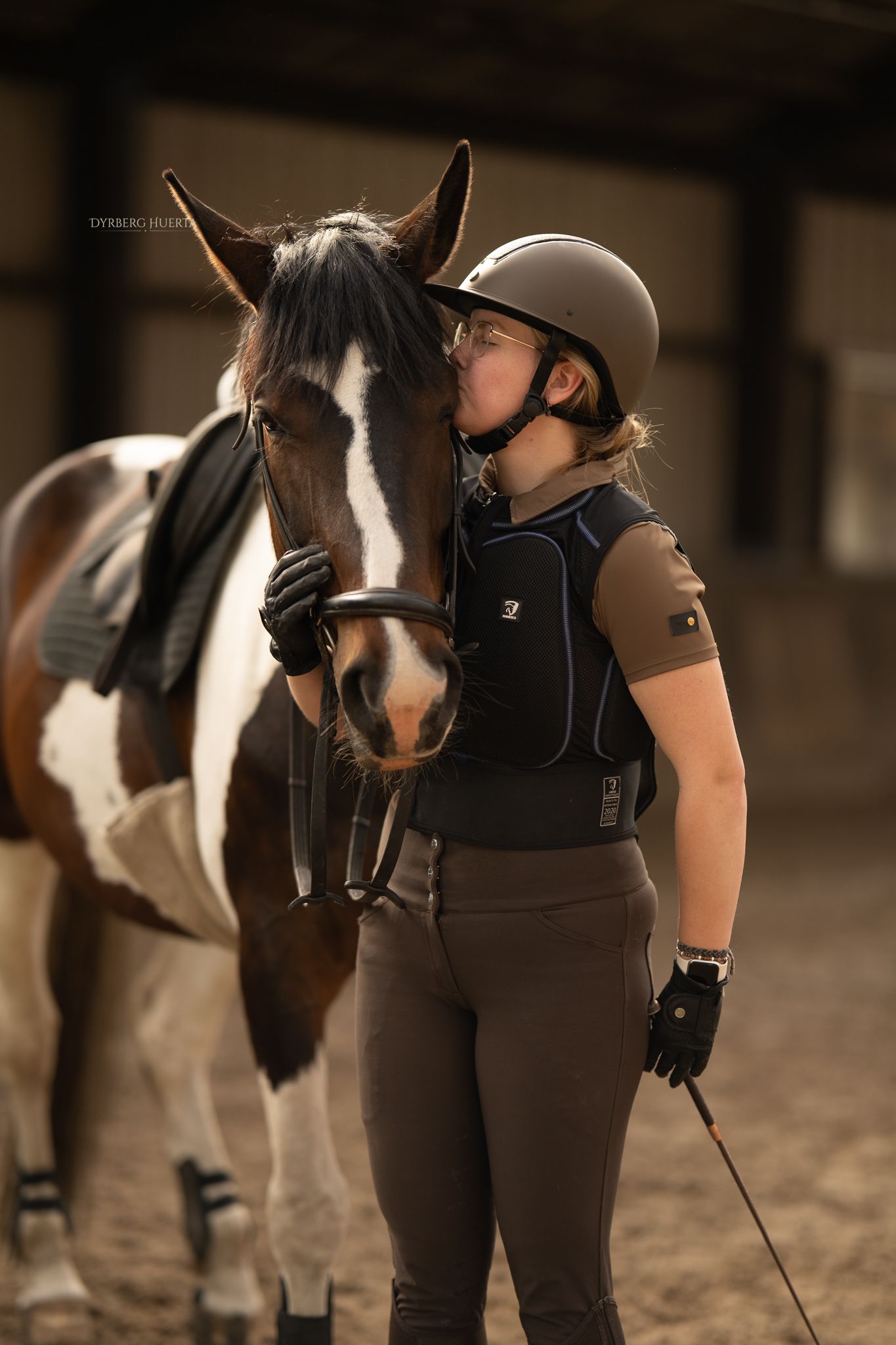 Young equestrian in helmet and vest shares a tender moment kissing her pinto horse in an indoor arena.