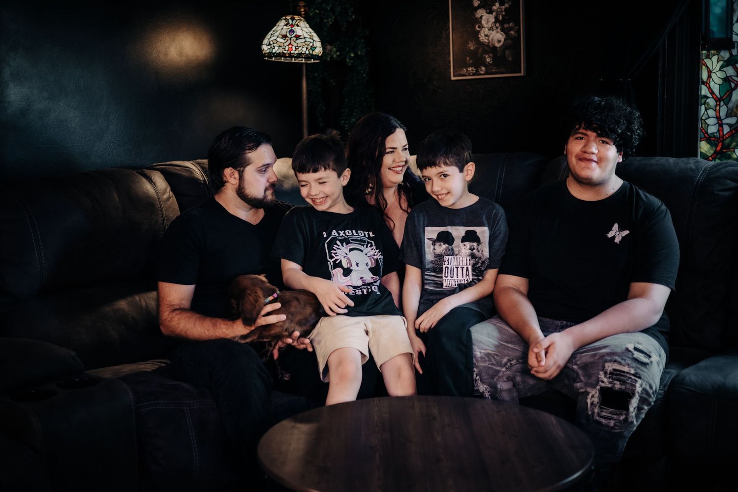 A woman with long dark hair in a black dress sits on a leather sofa with a young boy, sharing a tender moment.