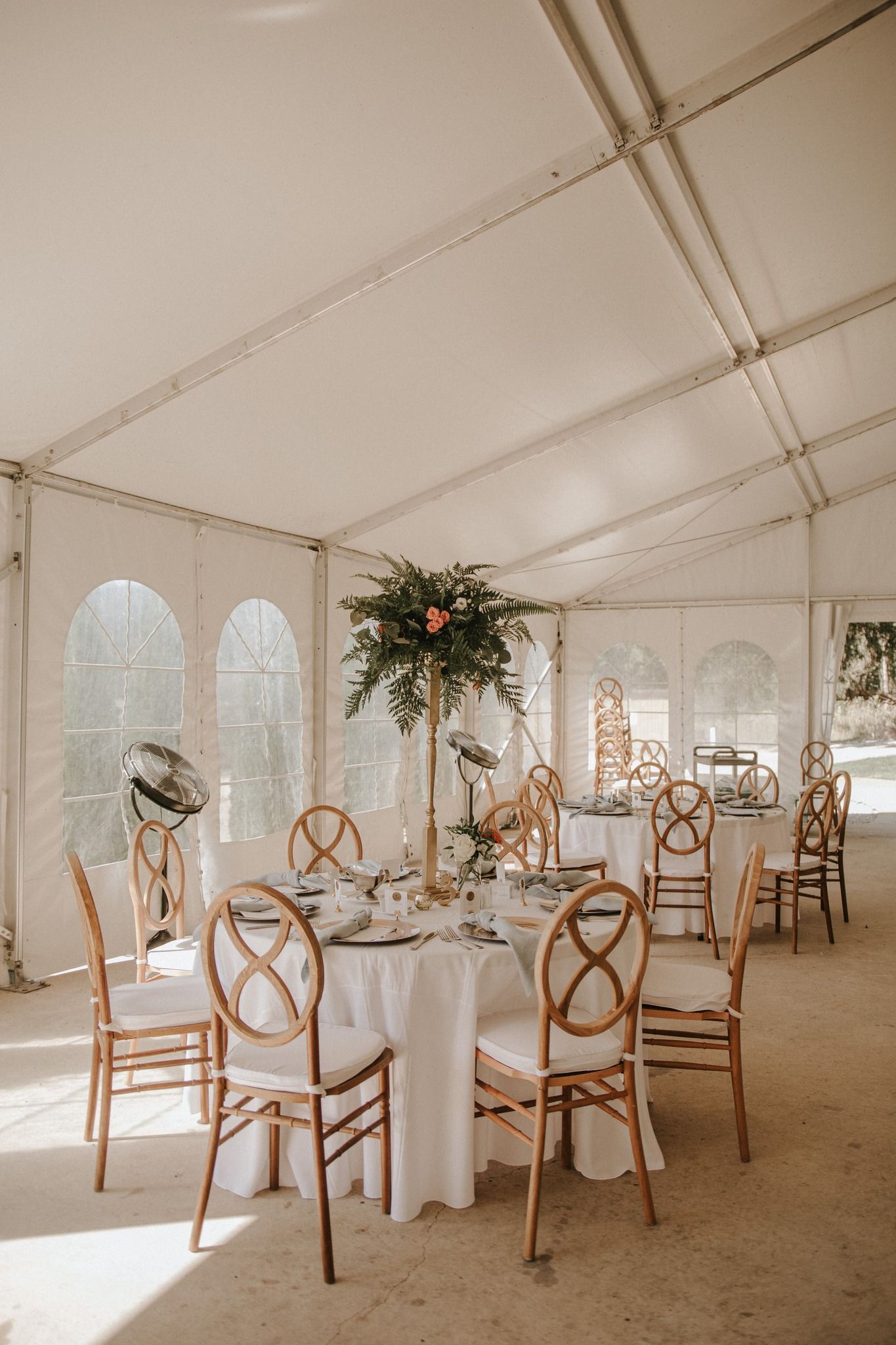 Elegant wedding tent reception with wooden cross-back chairs, white tablecloths, and tall floral centerpieces in natural light.