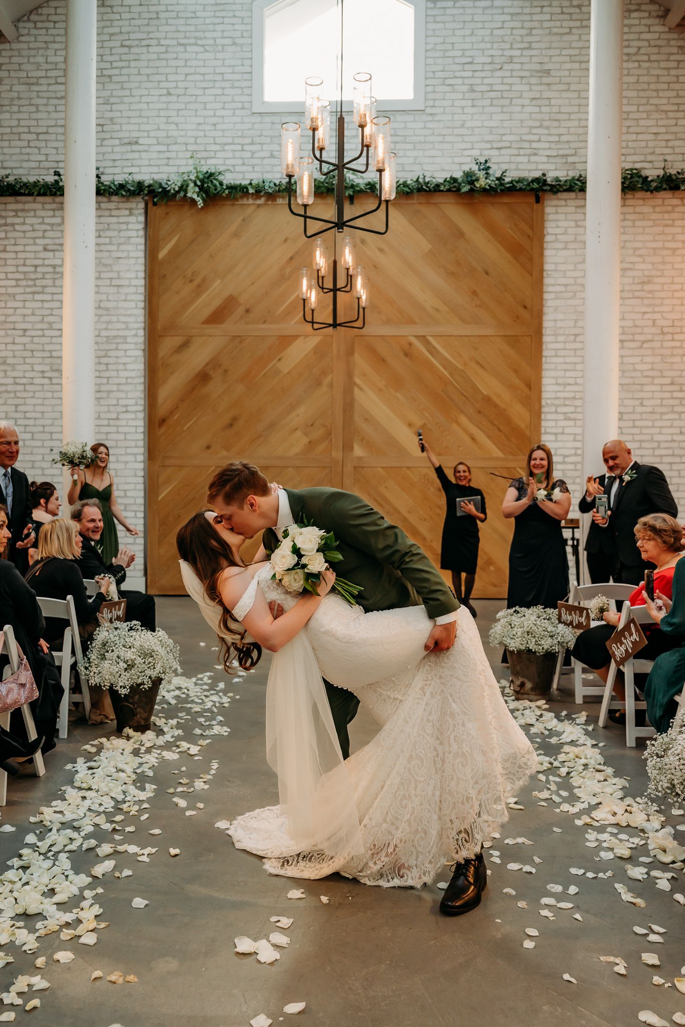 Bride in white gown and veil faces smiling groom in gray suit and cowboy hat during outdoor wedding ceremony.