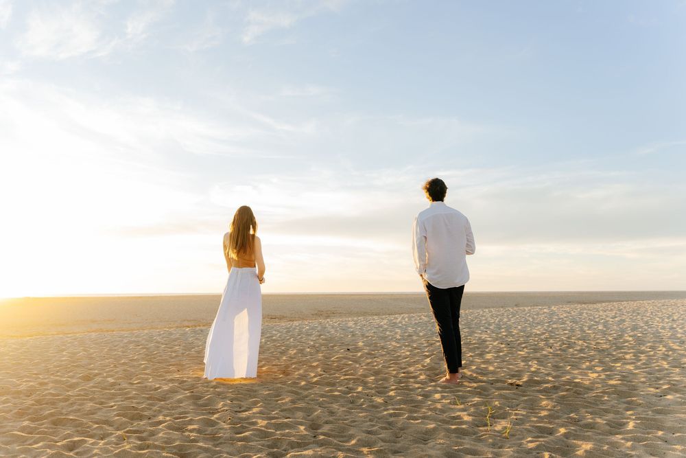 A couple in white attire walks along a sandy beach at golden sunset, viewed from behind.