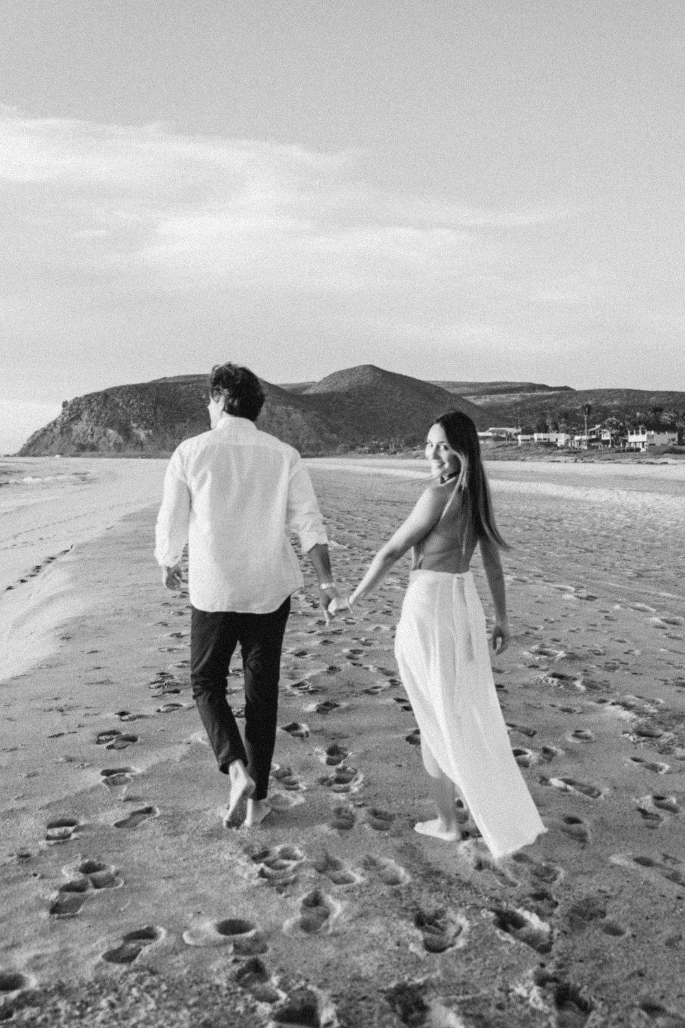 Black and white photo of a couple holding hands walking barefoot on a sandy beach with mountain backdrop.