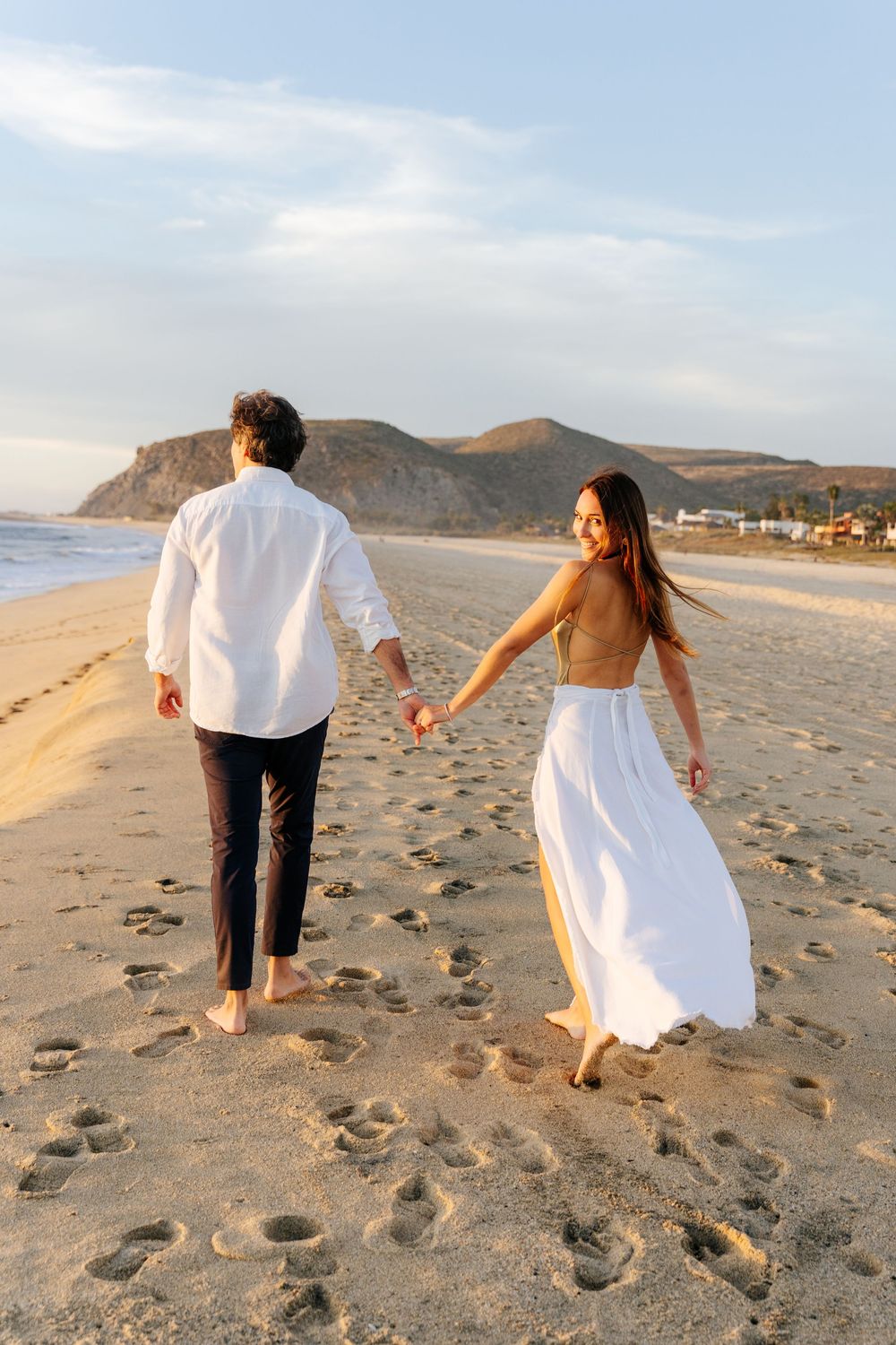 Couple holding hands walking barefoot on sandy beach at sunset with mountain backdrop.