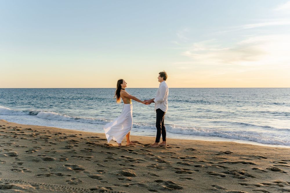 Couple holding hands on sandy beach at sunset, woman in white dress, romantic seaside engagement photo.