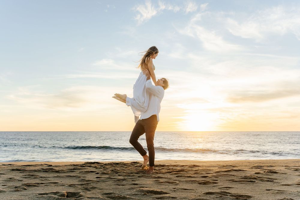 Romantic couple embracing at sunset on a sandy beach, man lifting woman in white dress by the ocean.