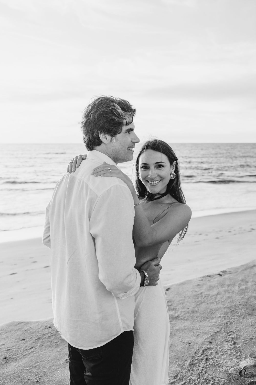 Black and white photo of a smiling couple embracing on a sandy beach with ocean waves in the background.