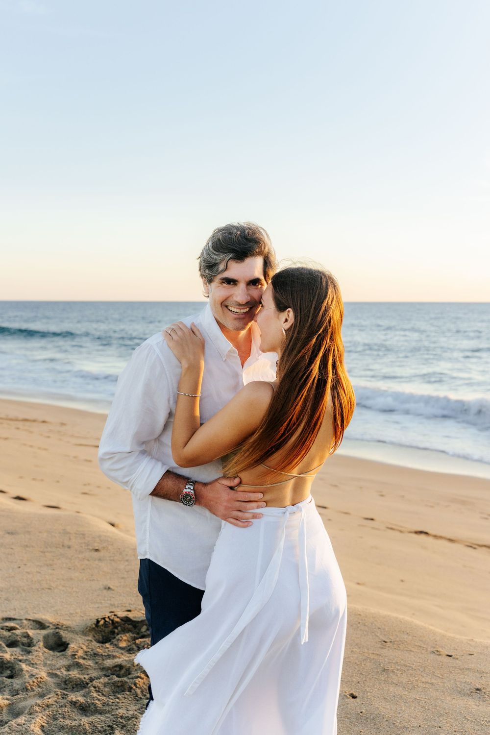 Happy couple embracing on a sunny beach at sunset, man smiling at camera, woman in white outfit.