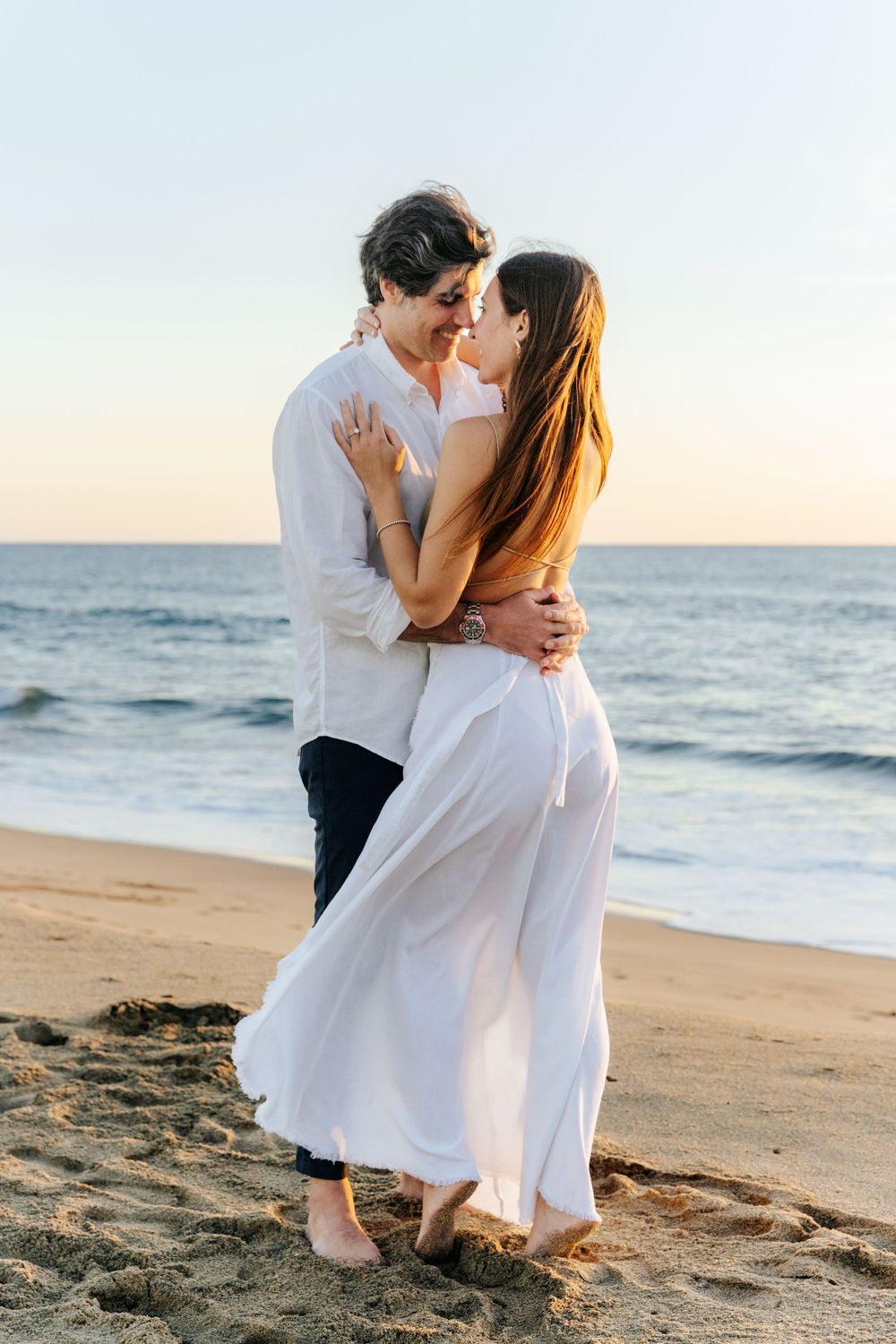 Couple sharing a romantic kiss on a sandy beach at sunset, woman in flowing white dress, ocean waves in background.