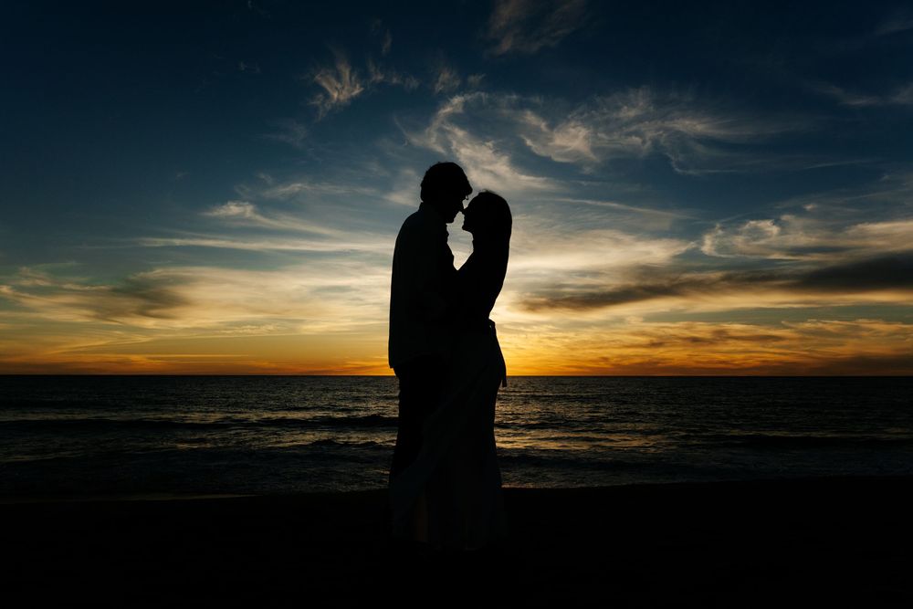 Romantic couple silhouette embracing at sunset on a beach with vibrant orange and blue sky over the ocean.