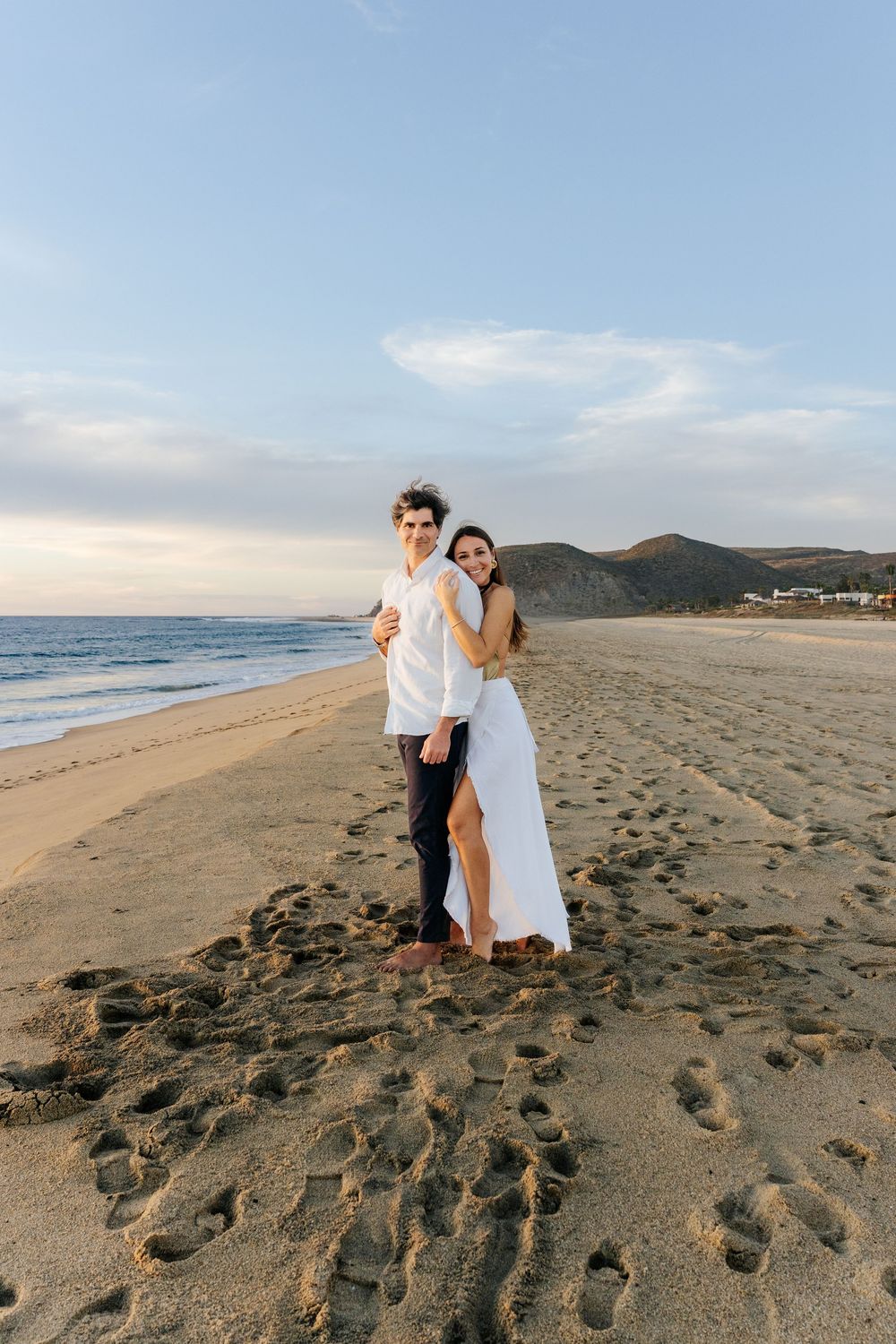 A happy couple embracing on a sunny beach with mountain views and footprints in the sand at golden hour.