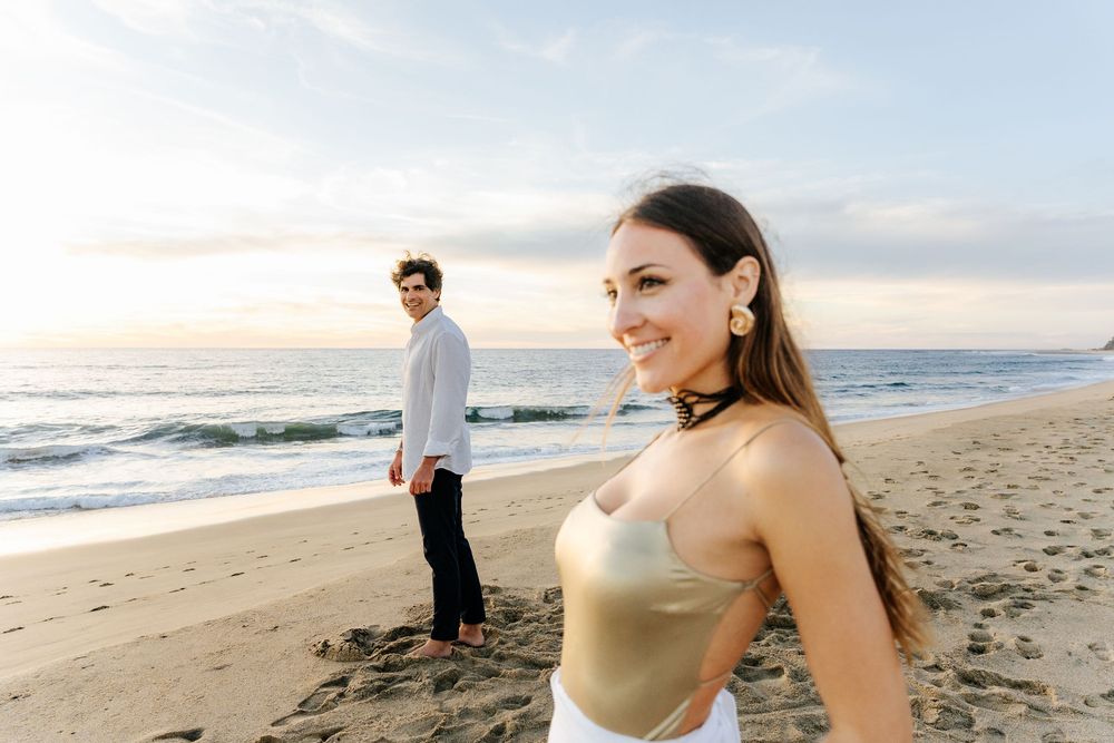 A smiling woman in a gold bodysuit poses on a sandy beach at sunset with a man standing near the ocean.