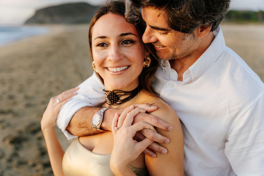 Smiling couple embracing on a beach, woman wearing gold jewelry and watch, man in white shirt at sunset.