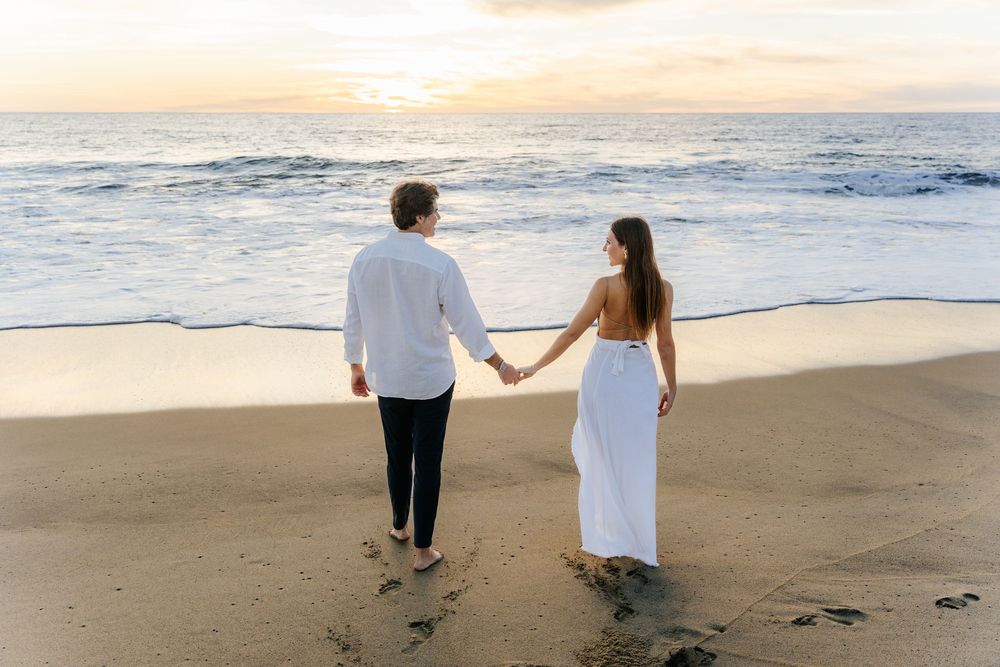 Couple holding hands walking toward ocean waves at sunset, woman in white dress, man in white shirt.
