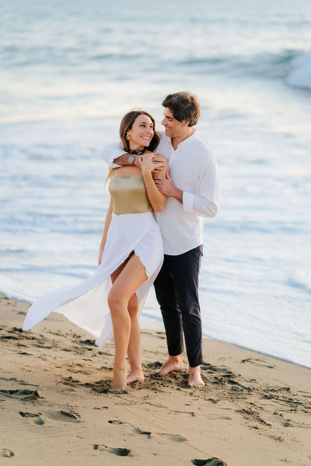 Romantic couple embracing on a sandy beach at sunset, woman in white dress, man in white shirt and black pants.