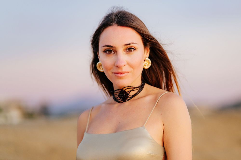 Young woman with dark hair wearing gold earrings and a choker necklace in a golden field at sunset.
