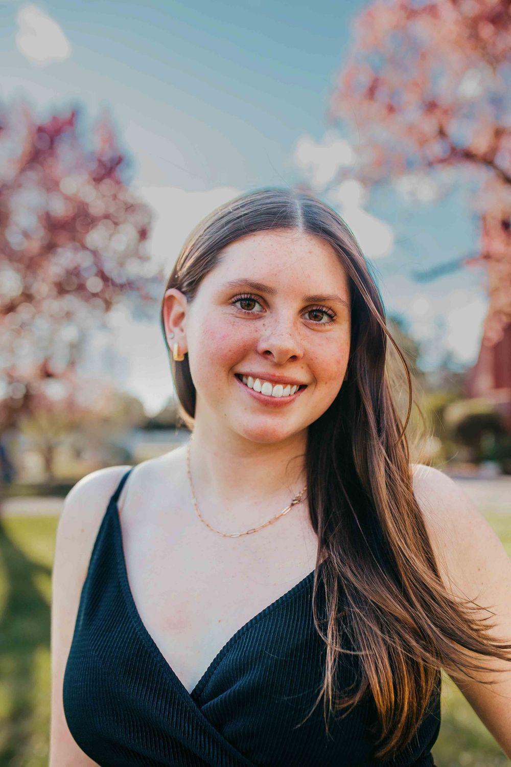 Smiling young woman with freckles and long brown hair wearing a black top and gold necklace outdoors near blooming trees.