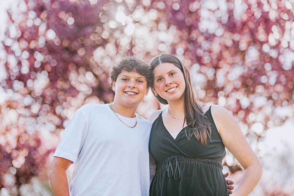 A smiling teen boy and woman pose together outdoors beneath blooming pink cherry blossom trees.