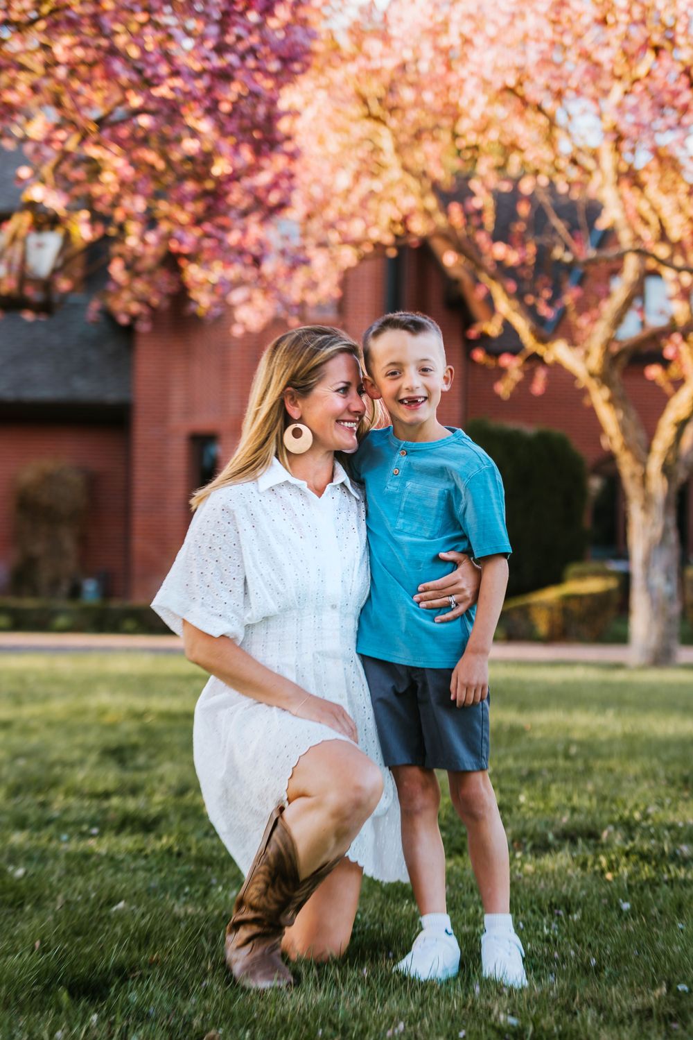 A woman in a white dress kneels beside a smiling boy in a teal shirt outdoors near blooming pink trees.